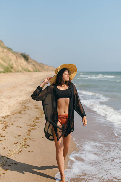 海の波で砂浜を歩いてリラックスした帽子を持つ美しい屈託のない女性。夏休みだ。薄い黒のシャツにスタイリッシュな若い女性にフィットし、熱帯の島で休暇を楽しむわら帽子 - 写真・画像