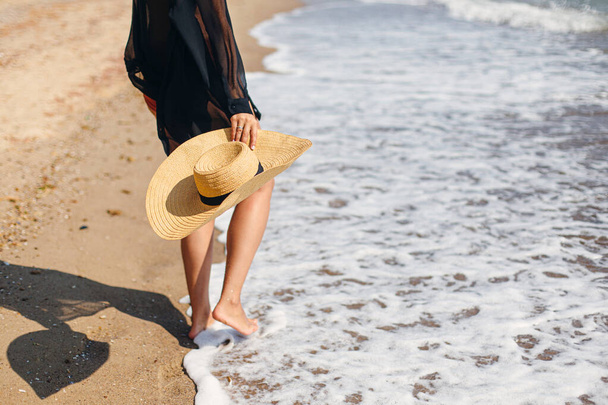 Summer vacation. Carefree woman holding hat and walking on sunny beach at sea waves, close up. Stylish young fit female with straw hat in hands relaxing on tropical shore. Space for text - Foto, Bild