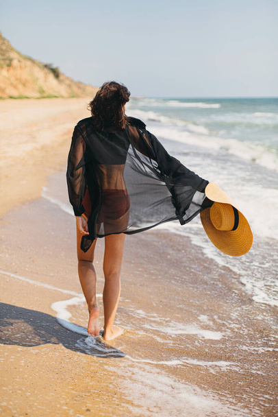 Mooie zorgeloze vrouw met hoed wandelen op zandstrand aan zee golven en ontspannen. Zomervakantie. Past stijlvol jong vrouwtje in licht zwart shirt en strohoed genieten van vakantie op tropisch eiland - Foto, afbeelding