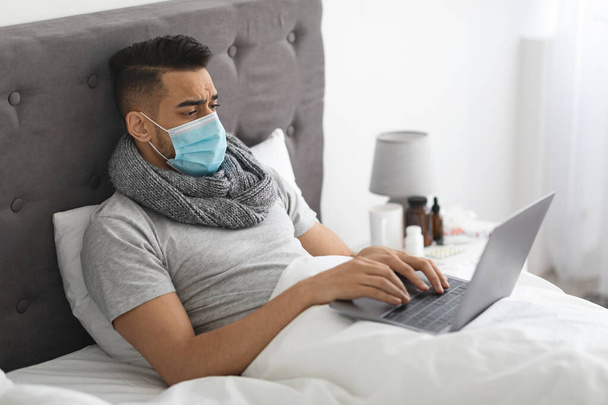 Enfermo hombre de Oriente Medio en máscara médica trabajando con el ordenador portátil en la cama - Foto, Imagen