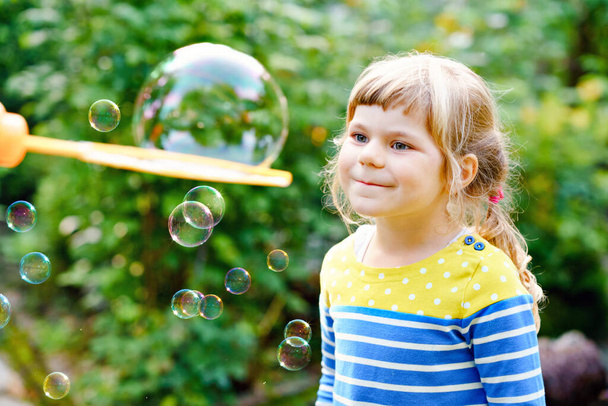 Boldog kis szőke óvodás lány szórakozik szappanbuborék fúvóval. Aranyos gyerek játszik a napos nyári napon. Boldog, aktív, vicces, egészséges gyereket. Tevékenység gyermekek számára. - Fotó, kép