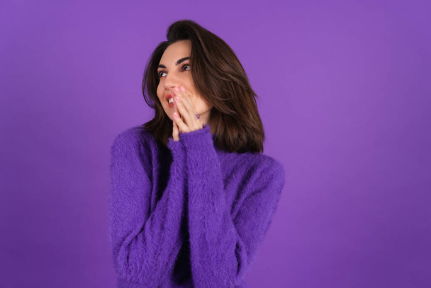 Young woman in a purple soft cozy sweater on the background is cute, in high spirits, laughs shyly, covering her mouth with her hands - Photo, image