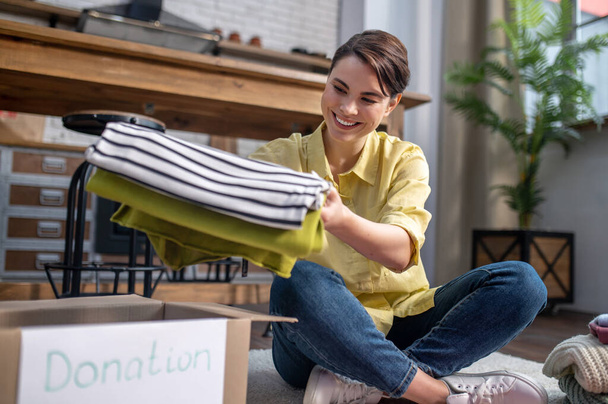 Joyous volunteer seated on the floor packing clothes for donation - Photo, image