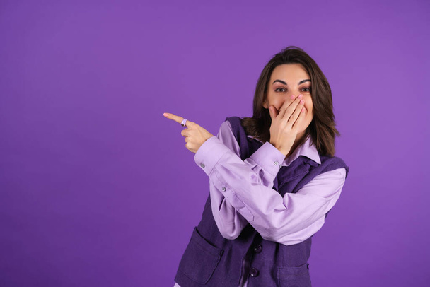 Young woman in a shirt and a vest on a purple background laughs loudly, covering her mouth with her hand, pointing her finger to the left - Foto, immagini