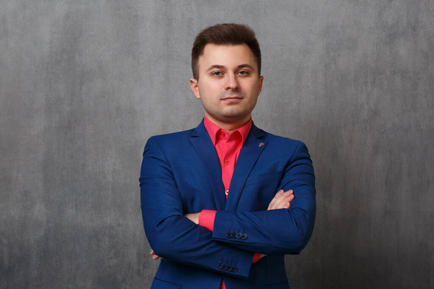 A handsome business man in a blue jacket and a bright pink shirt stands with his hands folded on his chest on a gray background. - Foto, afbeelding