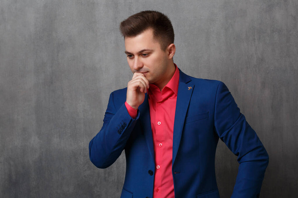 A handsome business man in a blue jacket and a bright pink shirt stands in thought with his hand to his chin on a gray background. - Photo, image