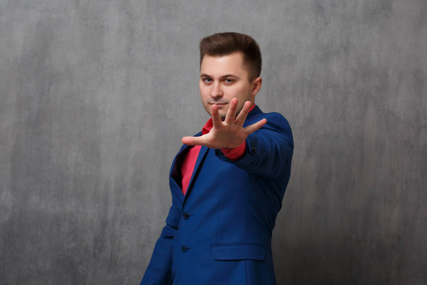 A young business man in a casual blue suit stretches out his hand in a blocking gesture against a gray background. - Foto, afbeelding