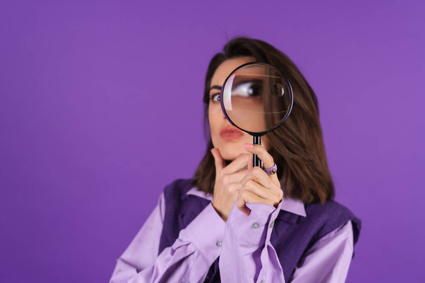 Young woman in shirt and vest on purple background having fun with magnifying glass in hand - Foto, afbeelding