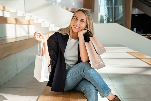 Candid portrait of a young woman in a white t-shirt, blue shirt and jeans in a mall holding shopping bags. Friendly layout concept - Photo, Image