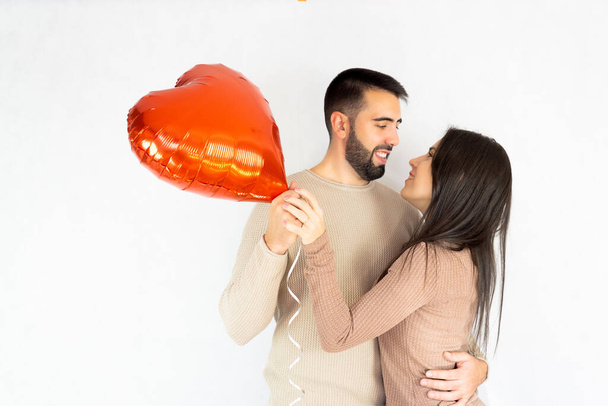 Couple in love looking happily at each other, holding a red heart-shaped balloon while embracing on a white background. - Fotó, kép
