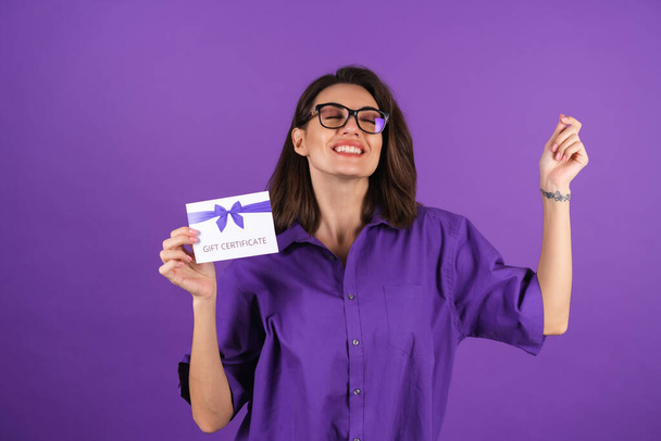 Young woman in a shirt on a purple background with a gift certificate smiling cheerfully, with makeup, lipstick on her lips and glasses, excited - Photo, image