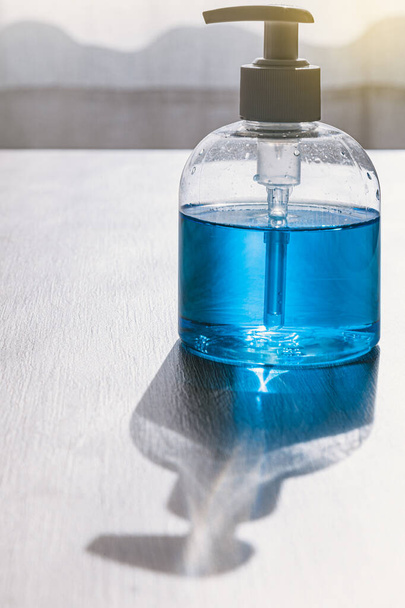 Op een houten tafel staat een fles met een dispenser van hydroalcoholische gel of blauwe zeep. Het licht komt intens van voren en werpt de schaduw op het tafelblad. Er is een venster op de achtergrond. - Foto, afbeelding