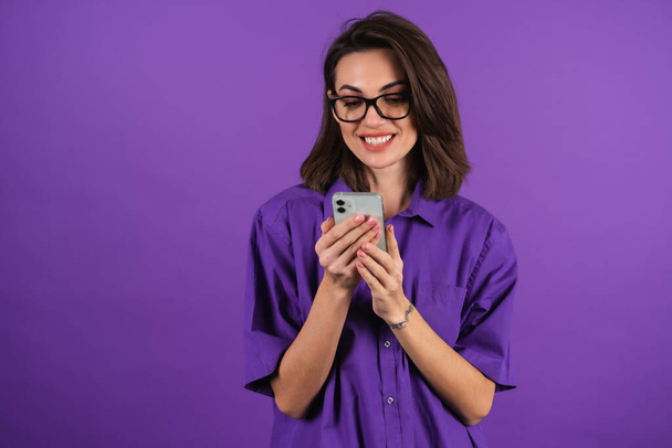 Mujer joven en una camisa y gafas sobre un fondo púrpura con un teléfono mira a la pantalla con una sonrisa alegre, leyendo un mensaje - Foto, imagen