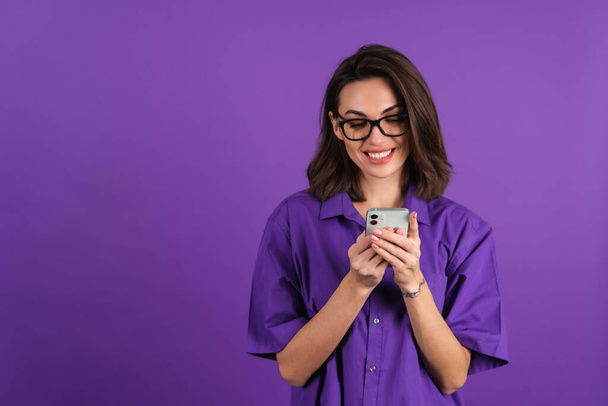 Young woman in a shirt and glasses on a purple background with a phone looks at the screen with a cheerful smile, reading a message - Fotoğraf, Görsel