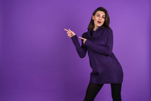 Young woman in a knitted warm dress and black tights on a purple background, beautiful figure, smiling cheerfully, pointing her finger to the left - Foto, Imagem