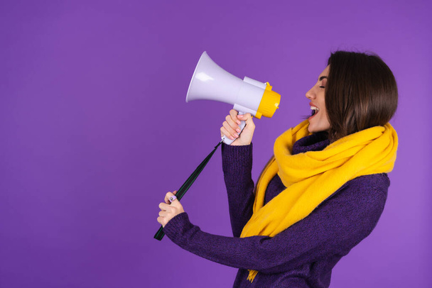 Young woman in a knitted dress and in a yellow scarf on a purple background cheerfully shouts into a megaphone, cheerful, announces a sale - Фото, изображение