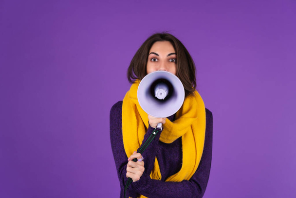 Young woman in a knitted dress and in a yellow scarf on a purple background cheerfully shouts into a megaphone, cheerful, announces a sale - Foto, afbeelding