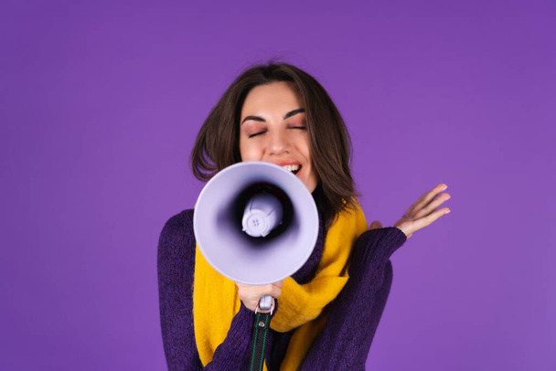 Young woman in a knitted dress and in a yellow scarf on a purple background cheerfully shouts into a megaphone, cheerful, announces a sale - Fotó, kép