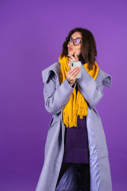 A young woman in a gray coat, a knitted dress and in a yellow scarf on a purple background, fashionable glasses, holds a phone and looks pensively to the side - Foto, afbeelding