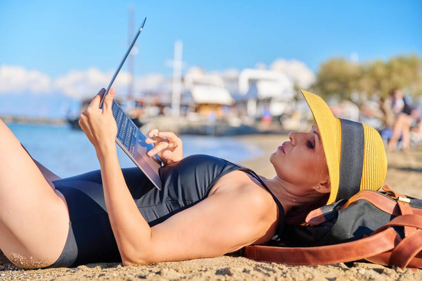 Mature woman sunbathing on the beach in hat with laptop - Foto, Imagen