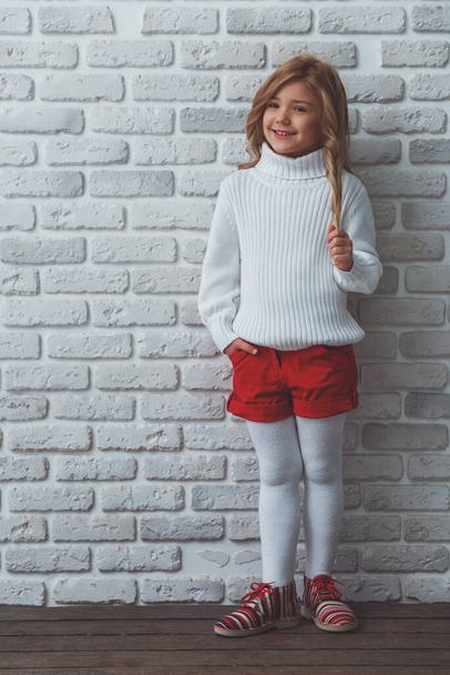Pretty little blonde girl looking in camera and smiling while standing against white brick wall - Photo, Image