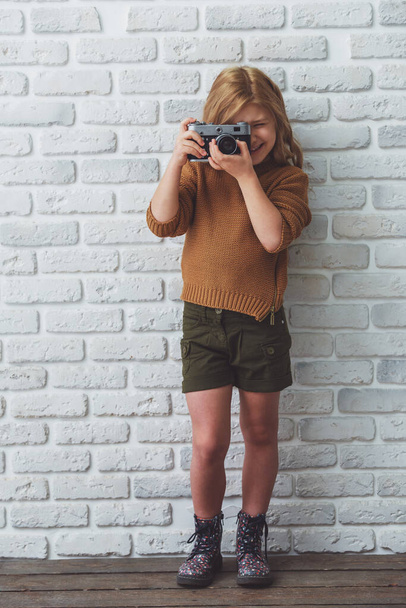 Pretty little blonde girl smiling and making a photo while standing against white brick wall - Zdjęcie, obraz