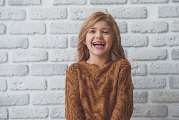 Portret van een mooie kleine blonde meisje in de camera kijken en lachen terwijl je tegen witte bakstenen muur - Foto, afbeelding