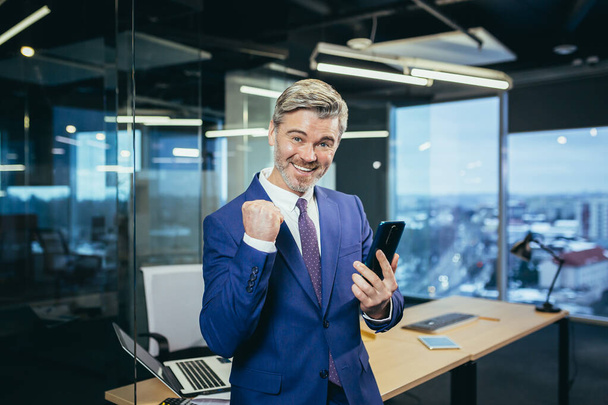 Happy boss rejoices in victory, businessman in modern office looks at camera and smiles, uses phone and app to buy shares online - 写真・画像