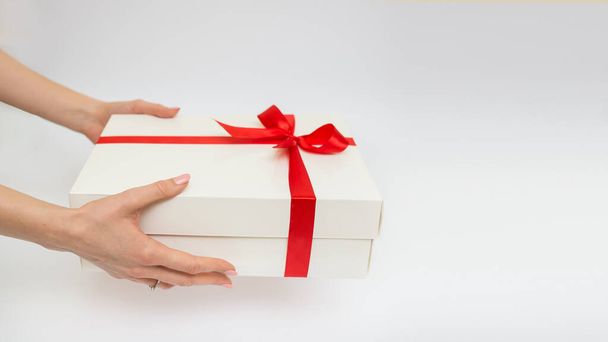 Женщины держат в руках подарочную коробку белого цвета с красной лентой, один предмет на белом фоне. Подарочный праздник. Праздничный фон. Вид сверху. Свободное место для текста - Фото, изображение