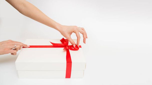 Manos femeninas sosteniendo una caja de regalo blanca con una cinta roja, un objeto sobre un fondo blanco. Vacaciones de regalo. Fondo festivo. Vista superior. Espacio libre para texto - Foto, imagen