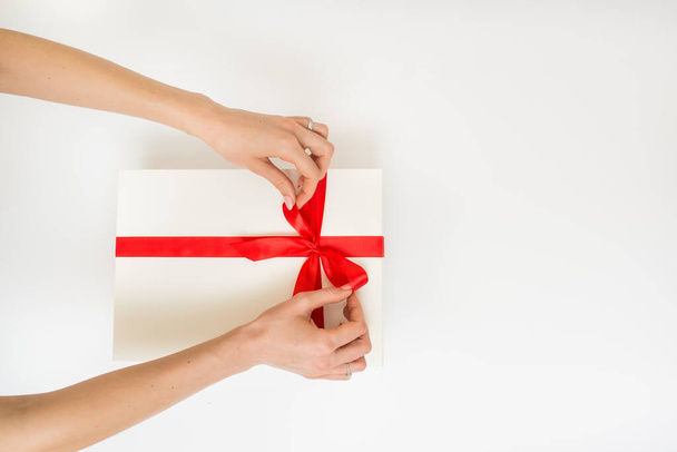 Manos femeninas sosteniendo una caja de regalo blanca con una cinta roja, un objeto sobre un fondo blanco. Vacaciones de regalo. Fondo festivo. Vista superior. Espacio libre para texto - Foto, imagen