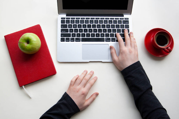 Muž pravou rukou na klávesnici notebooku a levou rukou na bílém stole s červeným notebookem a šálkem kávy, zelené jablko - Fotografie, Obrázek