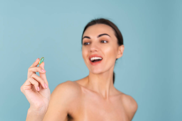 Γκρο πλαν πορτρέτο ομορφιάς μιας γυμνόστηθας γυναίκας με τέλειο δέρμα και φυσικό make-up, κρατώντας ένα συμπλήρωμα, βιταμίνη ωμέγα 3, ιχθυέλαιο, σε τυρκουάζ φόντο - Φωτογραφία, εικόνα