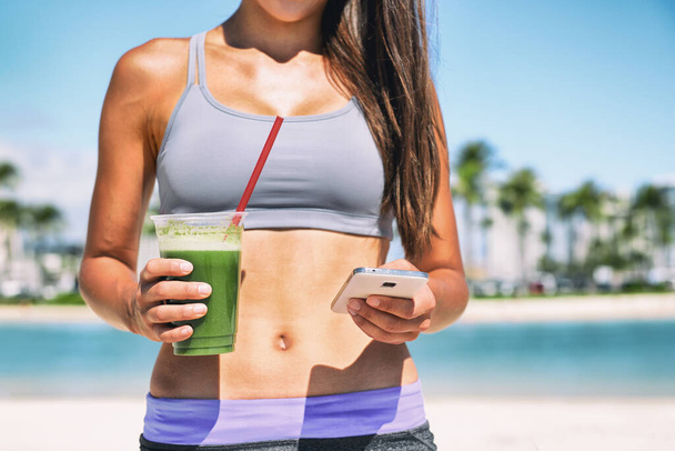 Healthy green smoothie juice drink online order on phone app fit athlete girl drinking detox cleanse morning breakfast on beach. Sport woman outside in summer sun with diet food - Foto, Imagen