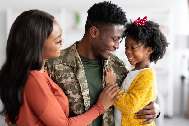 feliz negro soldado hombre abrazando con esposa y poco hija en casa - Foto, Imagen