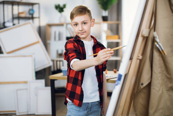 Caucasian boy standing in front of easel with brush in hands - Foto, imagen
