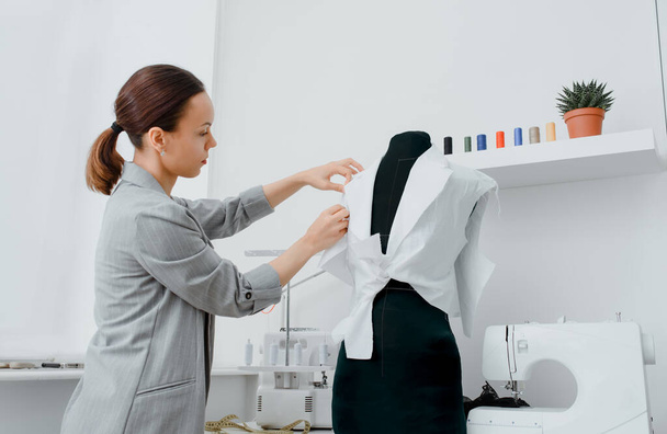 Η νεαρή ράφτρα κανονίζει ένα μελλοντικό πουκάμισο σε μια κούκλα στο μίνι εργαστήριό της. Στο τραπέζι είναι: overlock και ραπτομηχανή. Σχέδιο δημιουργικού σχεδιασμού. - Φωτογραφία, εικόνα
