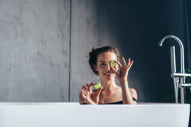 Woman sits in the bathtub and holds cucumber slices before applying them to her face. - Foto, Imagem