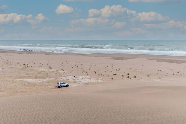 View from the top of a sand dune of a vehicle in the dunes. Behind more dunes, the sea and the cloudy sky - Fotoğraf, Görsel