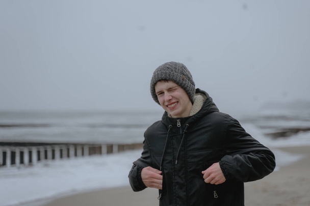 portrait of a teenager at the sea, a guy in a winter hat and a warm jacket squints against the strong wind, looks into the camera - Photo, image