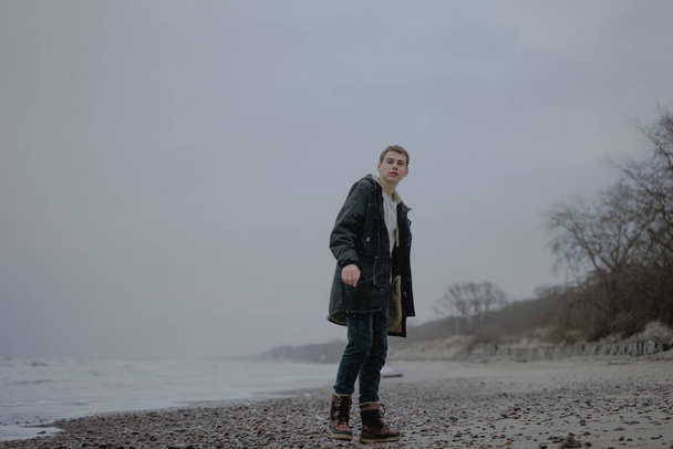 teenager stands on the seashore in a black jacket, looks at the camera, seascape cold weather - Foto, Bild