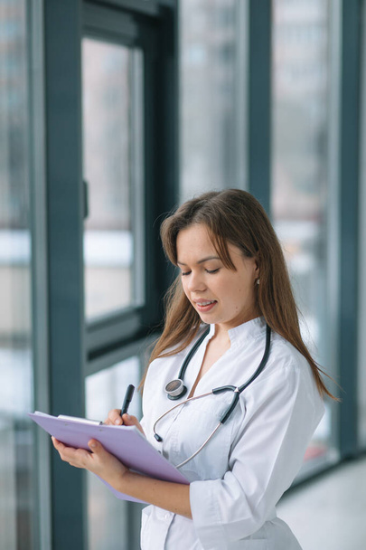 Young woman doctor wearing white medical coat and stethoscope writes a medical history in a notebook near the window - Фото, изображение
