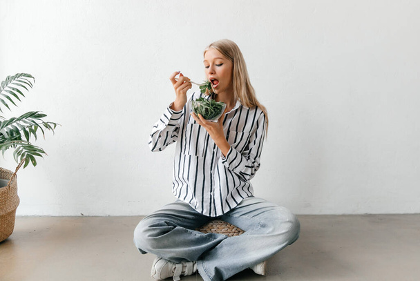 Koncepcja zdrowego stylu życia. Młoda blondynka siedząca na podłodze, jedząca sałatkę i uśmiechnięta - Zdjęcie, obraz
