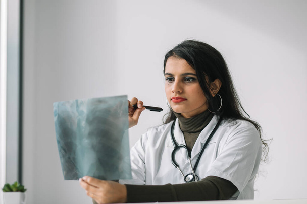 Indian female doctor in white coat with stethoscope examines x-ray and makes notes. Young indian medical student at the clinic in practice - Fotó, kép