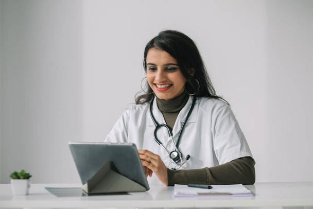 Medico donna indiana in un cappotto bianco con uno stetoscopio conduce una consultazione online nel suo ufficio - Foto, immagini