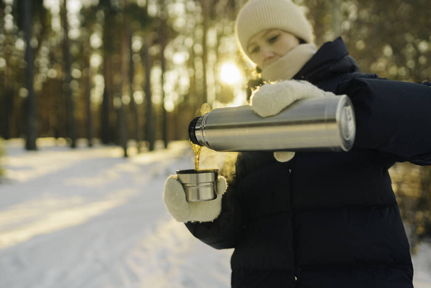 Woman traveler pours hot tea from a thermos into a mug, walking in snowy frozen winter forest. Adventure, travel, tourism and camping concept - 写真・画像