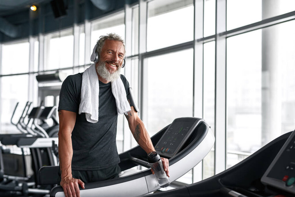 Cheerful grey-haired man with towel around neck, posing in gym - Zdjęcie, obraz