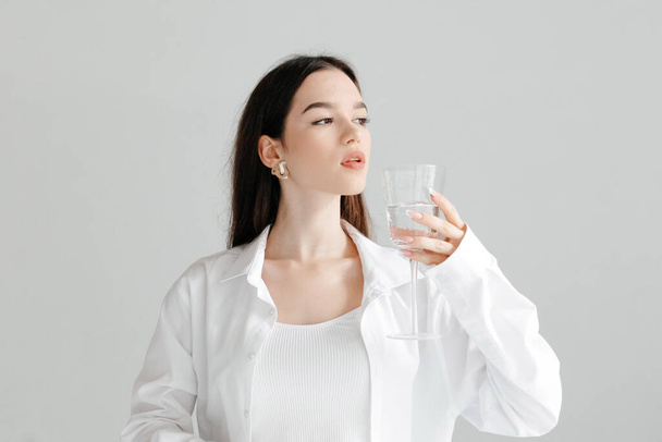 Krásná žena v bílé košili se sklenicí vody v ruce, na studio bílé pozadí. Zdravý životní styl, koncepce zdravotní péče. - Fotografie, Obrázek