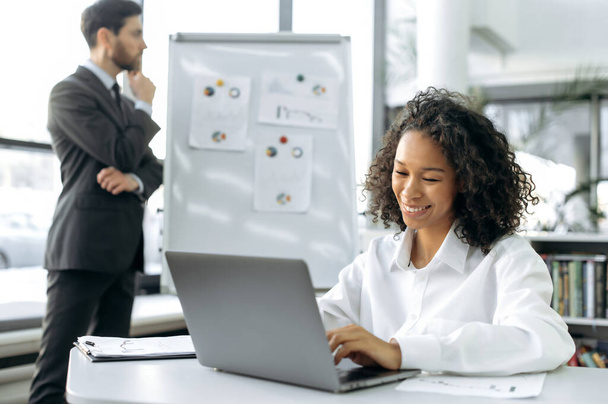 Positive young african american businesswoman, sales manager, sits in a modern office, working at laptop, smiles. On the background a male colleague ponders charts on a whiteboard, plans strategy - Фото, изображение