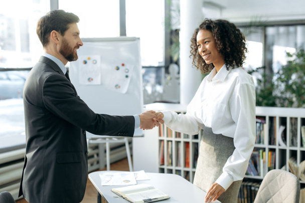 Handshake, agreement. Successful businessman and businesswoman, project partners, made a deal, cooperation, shake hands while standing in a modern office, smile at each other - Foto, afbeelding